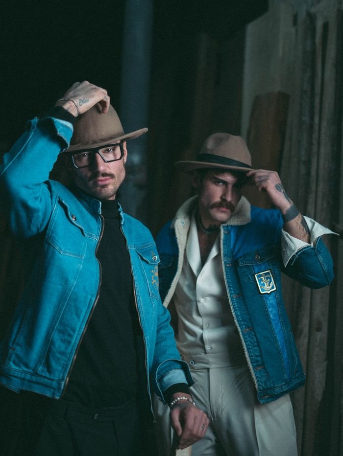 Two stylish men in denim jackets and hats posing indoors, exuding a modern fashion vibe.